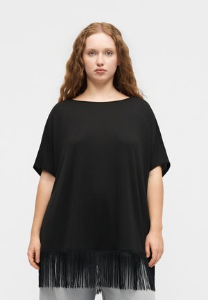 Γυναίκα με μακριά σγουρά μαλλιά, φοράει μια φαρδιά μαύρη μπλούζα με κοντά μανίκια και κρόσια στο τελείωμα, στέκεται μπροστά σε ένα απλό φωτεινό φόντο.