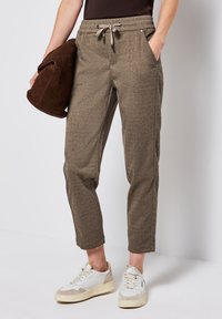 Persoon in een bruine broek met trekkoord, witte sneakers, die een gevouwen donkerbruine jas tegen een effen achtergrond vasthoudt.