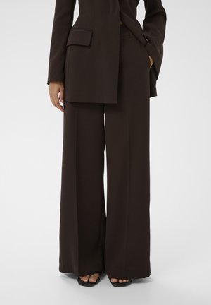 Braune, weit geschnittene Hosen und passender Blazer, getragen mit schwarzen Sandalen mit offenen Zehen, Hände entspannt an den Seiten, weißer Hintergrund.