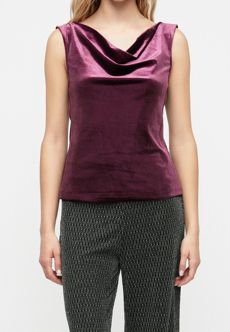 Femme portant un haut en velours violet sans manches à encolure drapée et un pantalon noir à motifs, debout devant un fond clair uni.
