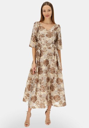 Kobieta w beżowej sukience midi z brązowymi kwiatowymi wzorami, z dekoltem w serek, rękawami do łokcia oraz beżowymi butami na obcasie z paskiem wokół kostki, stojąca na białym tle.