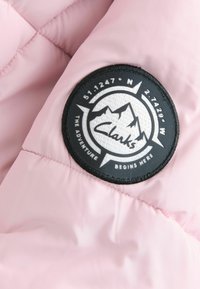 Tessuto trapuntato rosa chiaro con una toppa circolare nera caratterizzata da grafiche di montagne bianche e il testo "Clarks", posizionata nella zona della spalla.