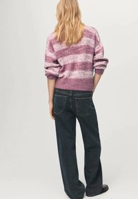 Donna bionda di spalle che indossa un maglione a righe rosa lavorato a maglia, jeans larghi scuri e scarpe nere su sfondo semplice.