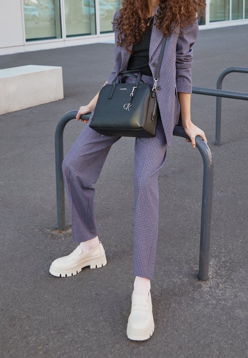 Personne portant un costume à carreaux violet et des chaussures blanches épaisses est assise sur une rambarde métallique en tenant un sac à main noir à l'extérieur.