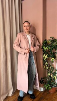 Person trägt einen langen rosa Mantel, weißes Hemd, blaue Jeans und schwarze Schuhe und steht drinnen in der Nähe von beigen Vorhängen und einer grünen Topfpflanze.