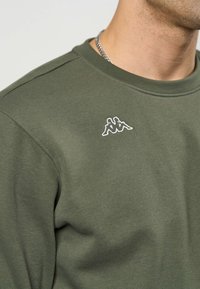 Bărbat purtând un hanorac verde măsliniu cu logo-ul alb Kappa pe piept și un lănțișor argintiu vizibil în jurul gâtului.