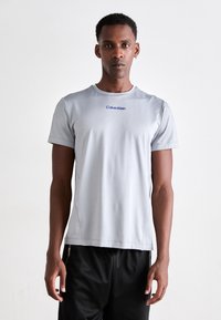 Tricou sportiv de culoare gri deschis, cu mâneci scurte, gât rotund și logo-ul albastru "Calvin Klein". Materialul pare neted, cu o textură subtilă.