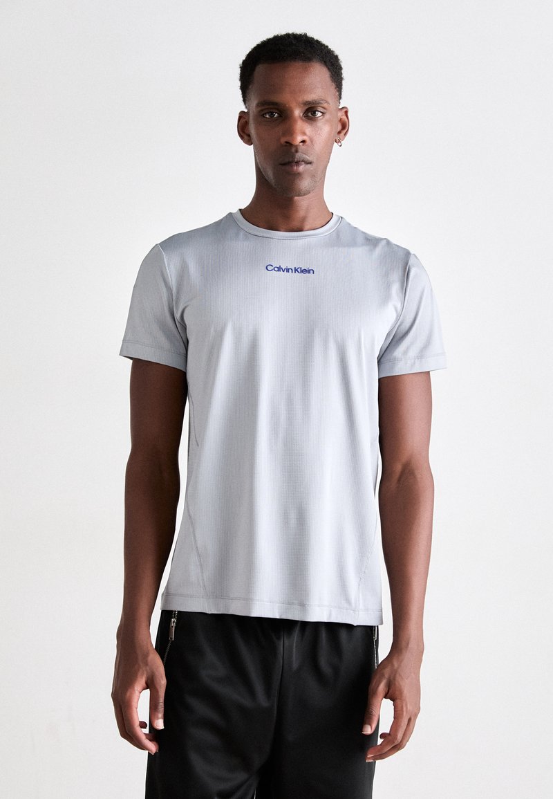 Világosszürke sportpóló rövid ujjal, kerek nyakkal és kék "Calvin Klein" logóval. Az anyag simának tűnik, finom textúrával.