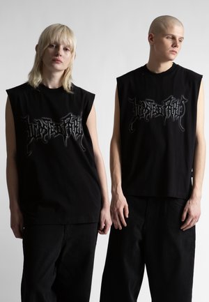 Δύο νέοι ενήλικες στέκονται πλάι-πλάι φορώντας μαύρα αμάνικα πουκάμισα με σκοτεινά γοτθικά γράμματα, ουδέτερες εκφράσεις, μπροστά σε απλό φωτεινό φόντο.