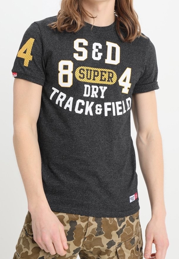 Person med en mørkegrå T-shirt med gule og hvide "S&D 84 SUPER DRY TRACK & FIELD" tekst, parret med brune camouflagebukser.