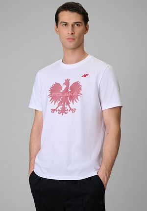 Młody mężczyzna stoi z rękami w kieszeniach, nosi białą koszulkę z czerwonym orłem i tekstem "Polska" z przodu na szarym tle.