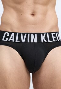 Crni pamučni bokserski gaćice s širokim elastičnim pojasom na kojem je u kontrastnim bijelim slovima ispisano "CALVIN KLEIN". Glatka tekstura i prilagođen dizajn.
