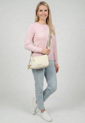 Žena stojící v světle růžovém svetru, světle modrých džínách, bílých teniskách a krémové kabelce přes rameno s přívěskem ve tvaru hvězdy.