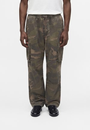 Homme portant un pantalon cargo camouflage ample avec poches latérales et des baskets noires sur fond blanc uni.