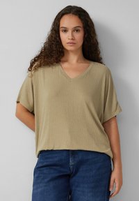 Vrouw met krullend haar, gekleed in een beige geribbelde V-hals losse top en blauwe denim jeans, tegen een effen lichtgrijze achtergrond.