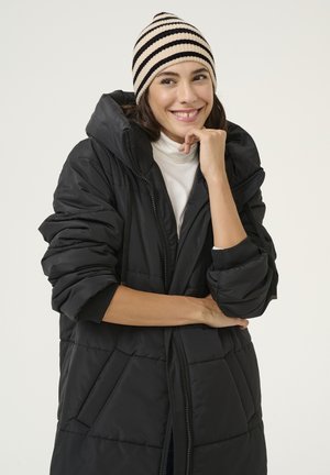 Cappotto imbottito nero con cappuccio, dotato di zip e tasche frontali. Indossato con un berretto lavorato a maglia a righe crema e nere.
