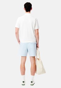 Camisa polo blanca, pantalones cortos a cuadros en azul claro, bolso de mano beige, calcetines blancos, zapatos con detalles en verde; visto desde atrás contra un fondo liso.