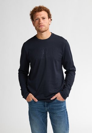 Marineblauwe longsleeve t-shirt met een ronde hals en subtiele logo-opdruk op de borst. Materiaal lijkt glad; gecombineerd met een blauwe spijkerbroek.
