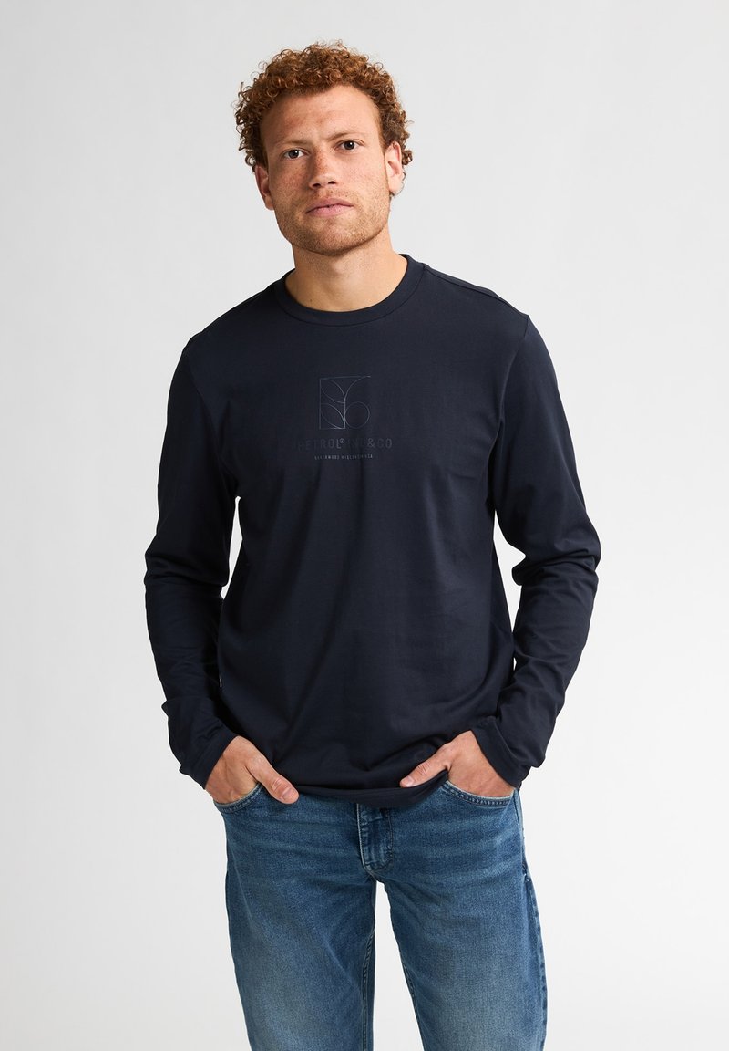 T-shirt a maniche lunghe blu navy con scollatura rotonda e discreto logo stampato sul petto. Il tessuto sembra liscio; abbinato a jeans blu.