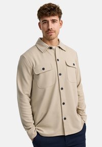 Camisa beige de botón con tejido texturizado y cuello, que presenta dos bolsillos en el pecho y botones negros. Diseño sencillo y ajuste recto.