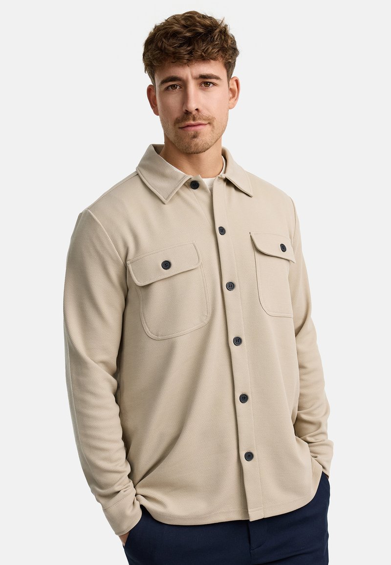 Camisa beige de botón con tejido texturizado y cuello, que presenta dos bolsillos en el pecho y botones negros. Diseño sencillo y ajuste recto.