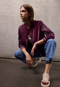 Bordo bomber bunda se pruhovanými manžetami, zipovou kapsou na rukávu, nošena přes bílý top a kombinována s roztrhanými modrými džíny a teniskami.