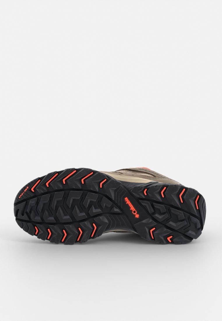 Chaussure de trail running avec une semelle extérieure en caoutchouc noir présentant de profondes rainures et des accents rouges ; tige en mesh beige avec un capuchon renforcé pour la durabilité.