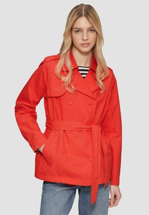 Trench-coat en coton rouge avec boutons croisés, taille ceinturée et poches latérales. Le mannequin porte un top rayé en dessous.