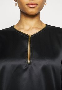 Haut en satin noir doté d'un col en V avec une fermeture éclair dorée, souligné par des surpiqûres dorées contrastantes et une texture lisse.