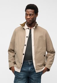 Νέος άνδρας με στρωμένο μπεζ μπουφάν, ανοιχτόχρωμο πουκάμισο, σκούρο μπλουζάκι και μπλε τζιν, στέκεται με τα χέρια στις τσέπες μπροστά σε λευκό φόντο.