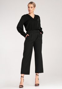 Czarna bluzka z długim rękawem, z dekoltem w kształcie litery V i plisowanym przodem, zestawiona z spodniami o wysokim stanie i szerokich nogawkach. Obie wykonane z gładkiego materiału.