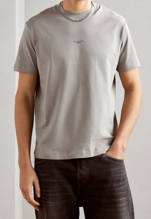 Homme portant un t-shirt gris clair à manches courtes Marc O’Polo Denim et un jean foncé, avec une chaîne en argent autour du cou et la main dans la poche.