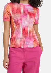 Top de manga corta en un tejido ligero y texturizado que presenta patrones geométricos en rosa, naranja y blanco, combinado con pantalones vibrantes en rosa.