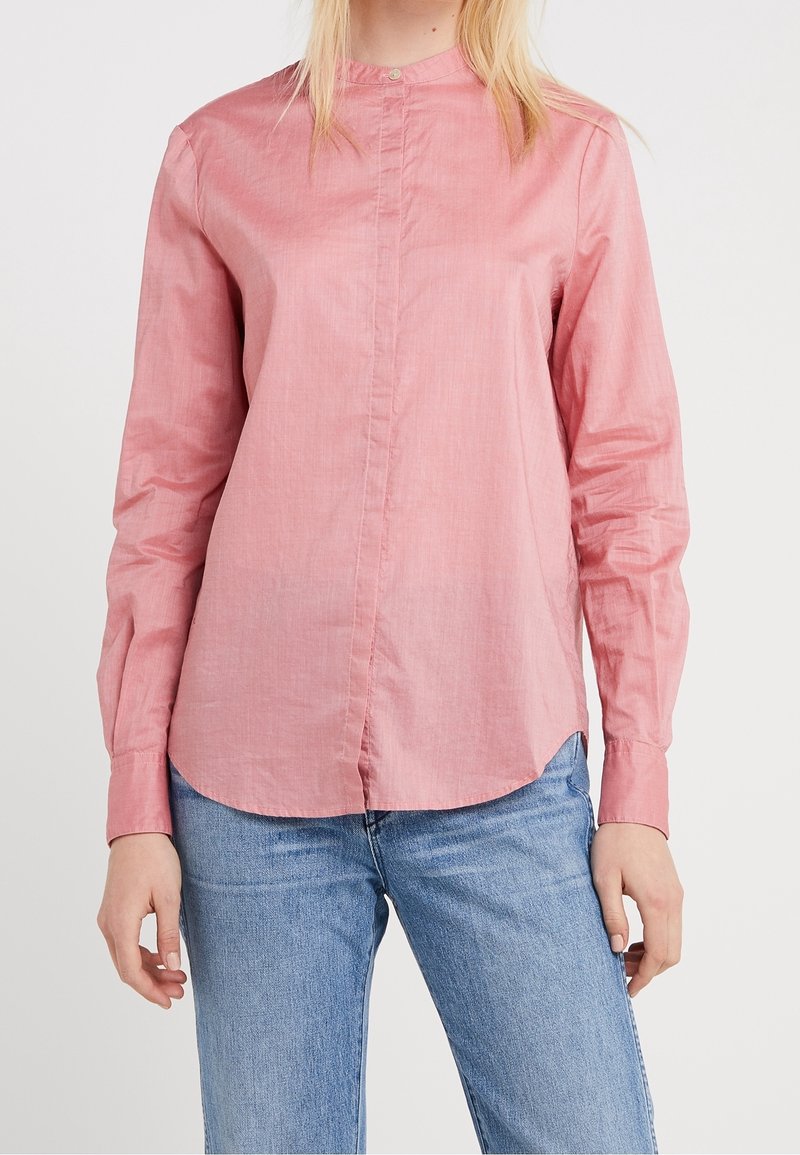 Femme portant une chemise rose à manches longues boutonnée et un jean bleu, debout devant un fond blanc uni.