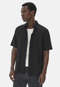 Chemise noire à manches courtes avec un tissu texturé, dotée d'un col et d'une coupe décontractée, portée par-dessus un débardeur blanc.