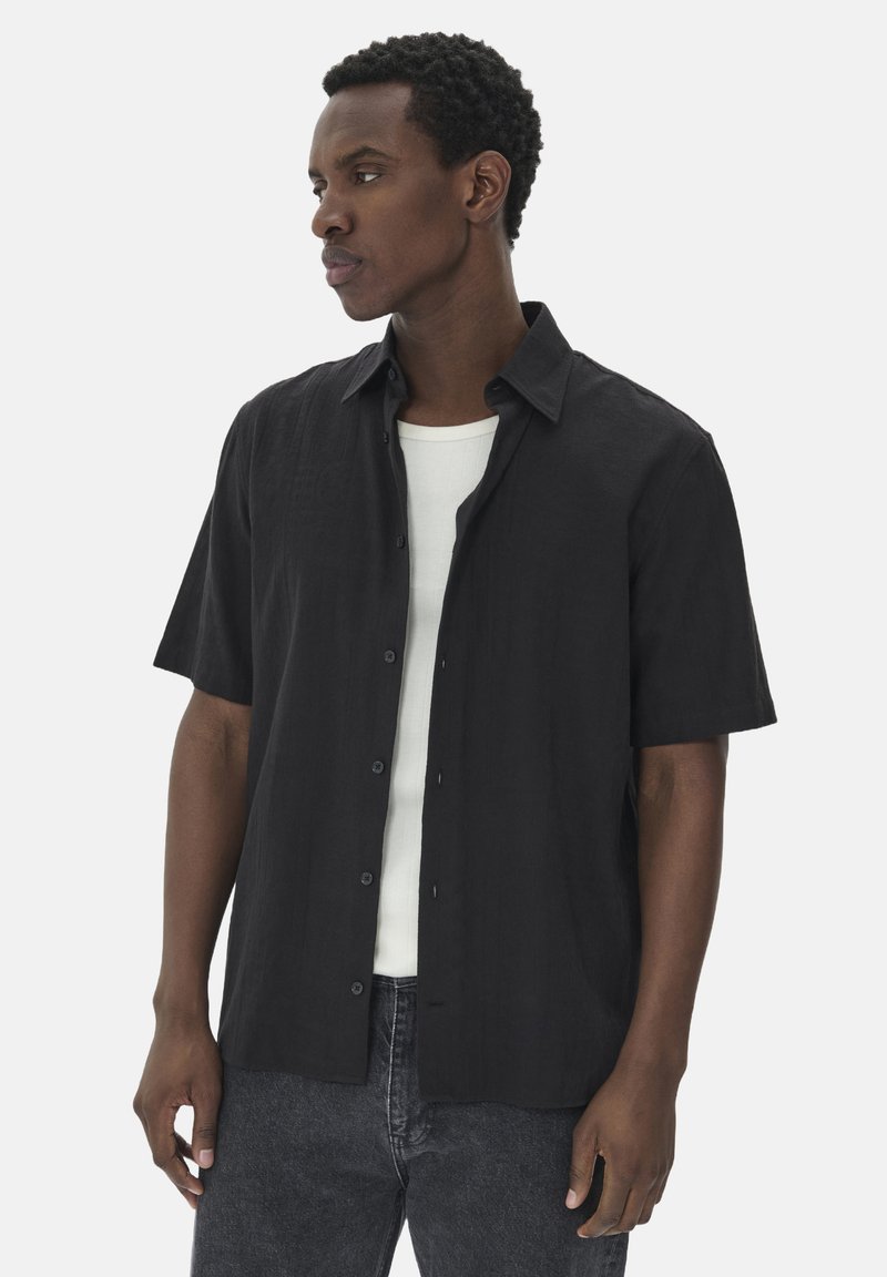 Chemise noire à manches courtes avec un tissu texturé, dotée d'un col et d'une coupe décontractée, portée par-dessus un débardeur blanc.