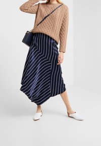 Beżowy sweter z dzianiny o teksturowanym wzorze, w połączeniu z granatową satynową spódnicą z diagonalnymi białymi paskami. Model ma na sobie białe buty wsuwane.