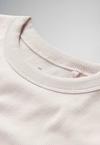 Detailný záber na golier svetlobéžového rebrovaného trička s okrúhlym výstrihom, na ktorom sú viditeľné švy a malá textilná slučka vo vnútri krku.