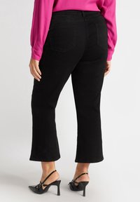 Schwarze hochgeschnittene Hose mit geradem Bein, strukturiertem Stoff und zwei Gesäßtaschen. Kombiniert mit schwarzen Absatzschuhen.