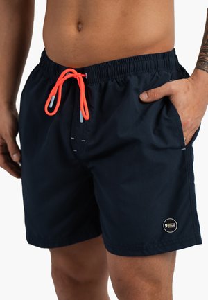 Homme portant un short de bain bleu marine avec un cordon orange fluo, main droite dans la poche, tatouage visible sur l’avant-bras gauche.