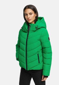 Femme portant une veste matelassée verte vif avec une capuche et une fermeture éclair noire, se tenant avec les mains dans les poches devant un fond uni.