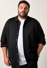 Giacca camicia nera con colletto, realizzata in tessuto texturizzato. Presenta due tasche frontali e una maglietta bianca a strati sotto.