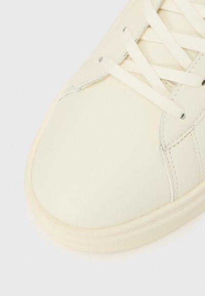 Ténis branco em pele focado na área dos dedos, com atacadores planos, textura lisa e biqueira arredondada. Detalhes subtis de costura são visíveis.