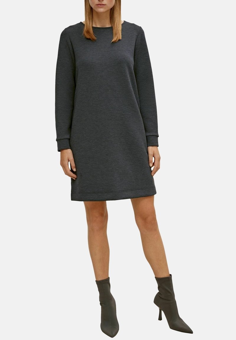 Femme portant une robe pull gris foncé à manches longues et des bottines assorties, debout devant un fond clair uni.