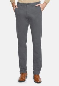 Hombre con pantalones rectos grises y zapatos de vestir de cuero marrón, con las manos en los bolsillos, de pie sobre un fondo blanco.