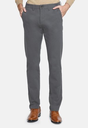 Hombre con pantalones rectos grises y zapatos de vestir de cuero marrón, con las manos en los bolsillos, de pie sobre un fondo blanco.