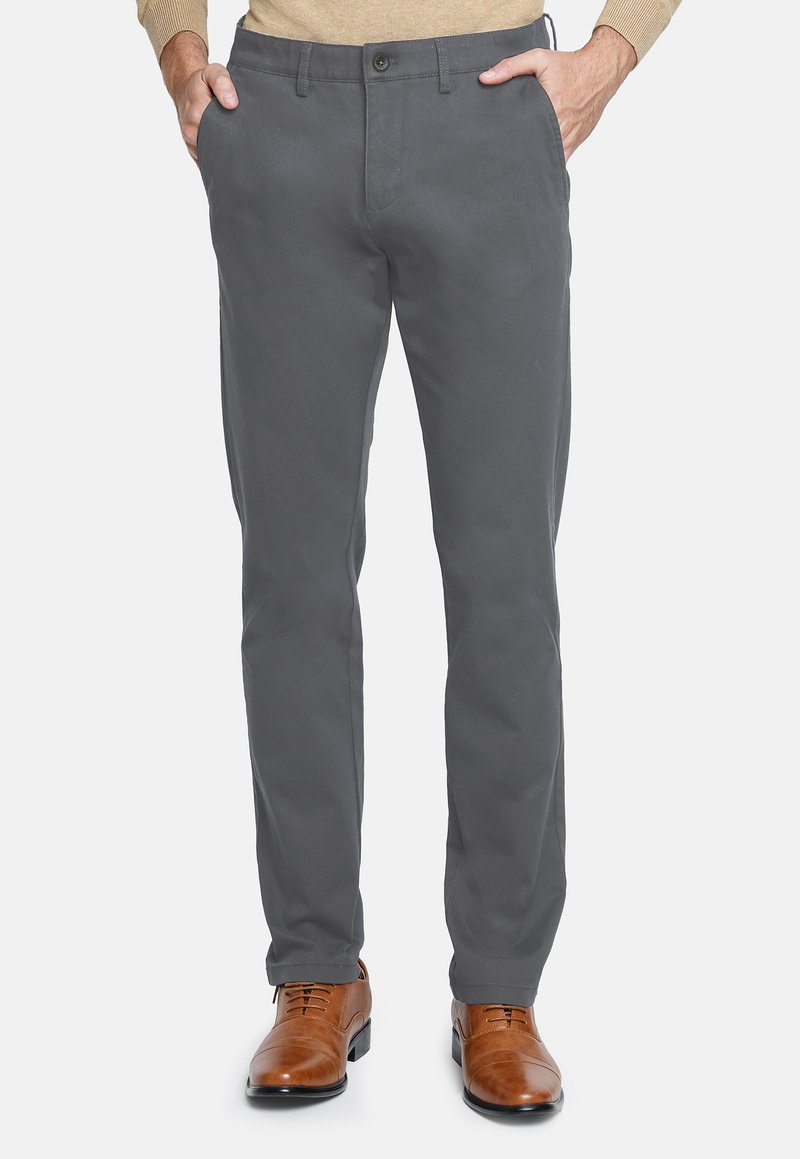 Hombre con pantalones rectos grises y zapatos de vestir de cuero marrón, con las manos en los bolsillos, de pie sobre un fondo blanco.