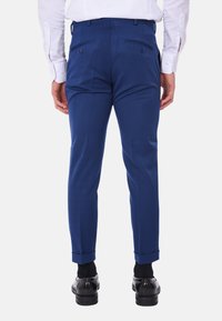 Pantaloni su misura blu con una texture liscia, dotati di tasche posteriori con bottoni e risvolti, indossati con scarpe nere e una camicia bianca.