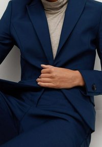 Costume bleu marine ajusté avec une veste cintrée présentant des revers crantés et une fermeture à un bouton noir, porté sur un col roulé gris clair.