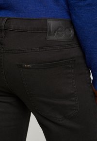 Närbild av svarta Lee-jeans bakficka och midjeband som bärs med en blå stickad tröja.